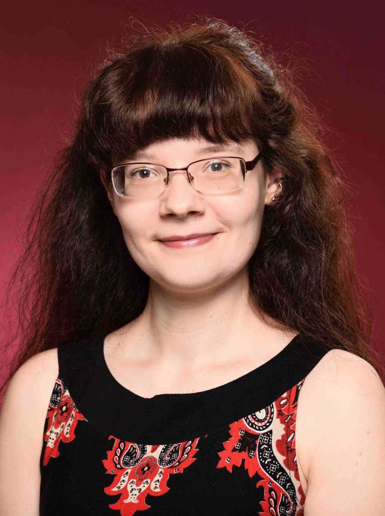 offizielles Pressefoto der Autorin Alicia Hartung vor einem roten Hintergrund im Profil; lange, braune Haare, dicker Pony, Brille